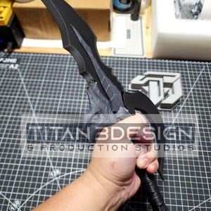 K&ouml;nnte beinhalten: Ein schwarzes und graues Dolch mit einer strukturierten Klinge und einem schwarzen Griff. Der Dolch wird von einer Hand gehalten. Der Text "TITAN3DESIGN & PRODUCTION STUDIOS" ist im Bild sichtbar.