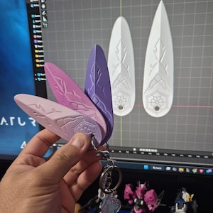 May include: A keyring with three shield-shaped charms in pink and purple gradients. The charms are attached to a silver keyring. Two white shield designs are visible in the background. The charms have a decorative design.