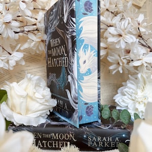 May include: Books titled "When the Moon Hatched" by Sarah A. Parker. The covers have a dark design with white and teal illustrations of a dragon and feathers. White flowers are arranged around the books, creating a decorative display.