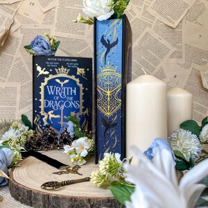 Op de afbeelding: Een marineblauwe boekvaas met gouden details en een draak-silhouet, en een boek met de titel "Wrath of the Dragons". De scène bevat kaarsen, bloemen en een sleutel op een houten schijf, wat een fantasiethema creëert.