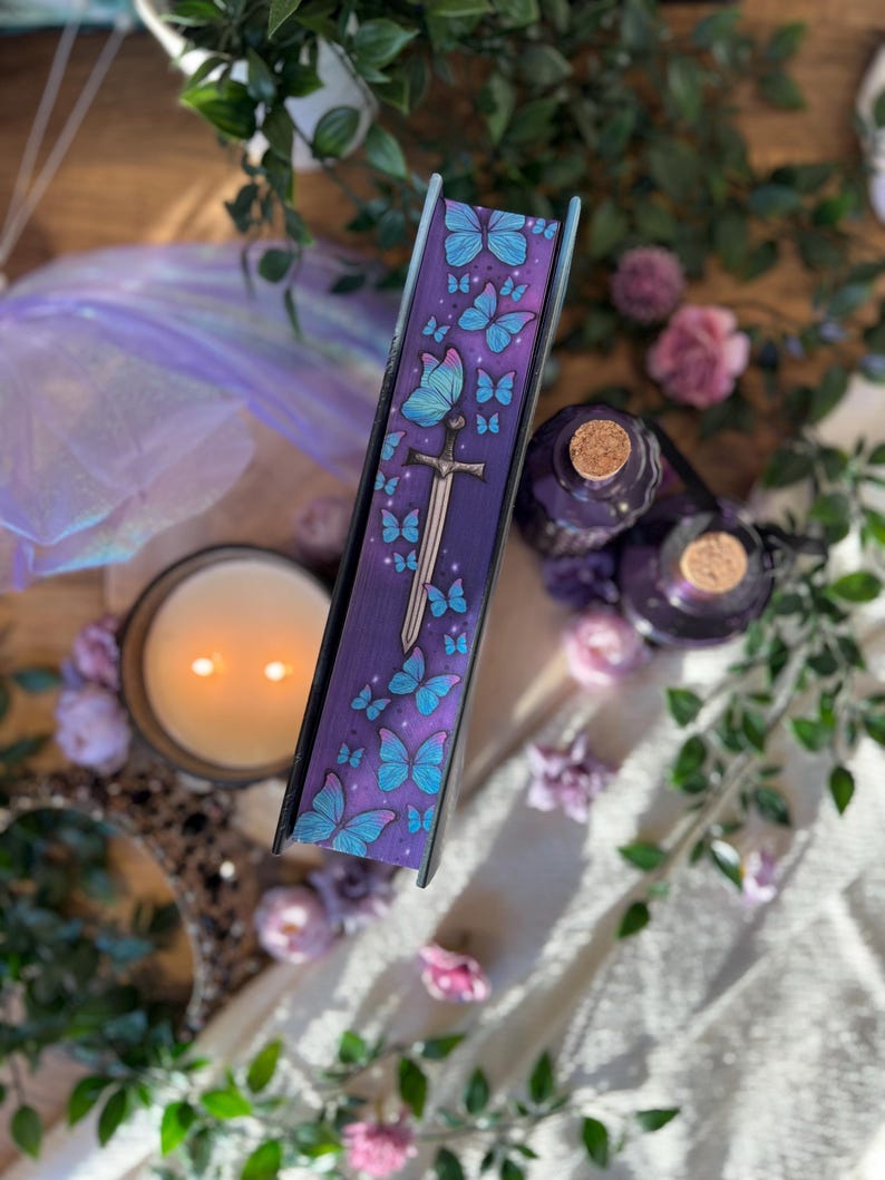 Puede incluir: Un libro morado con un dise&ntilde;o de mariposa y espada. El lomo del libro presenta mariposas azules y una espada plateada. El libro est&aacute; rodeado de velas, flores y botellas decorativas, creando una est&eacute;tica caprichosa.