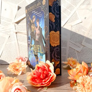 May include: A hardcover book with a dark blue cover and gold lettering. The title of the book is "Cruel Prince" and the author is Sophie Clark. The cover features a dark blue background with a gold snake and a gold crown. The book is surrounded by pink and orange flowers.