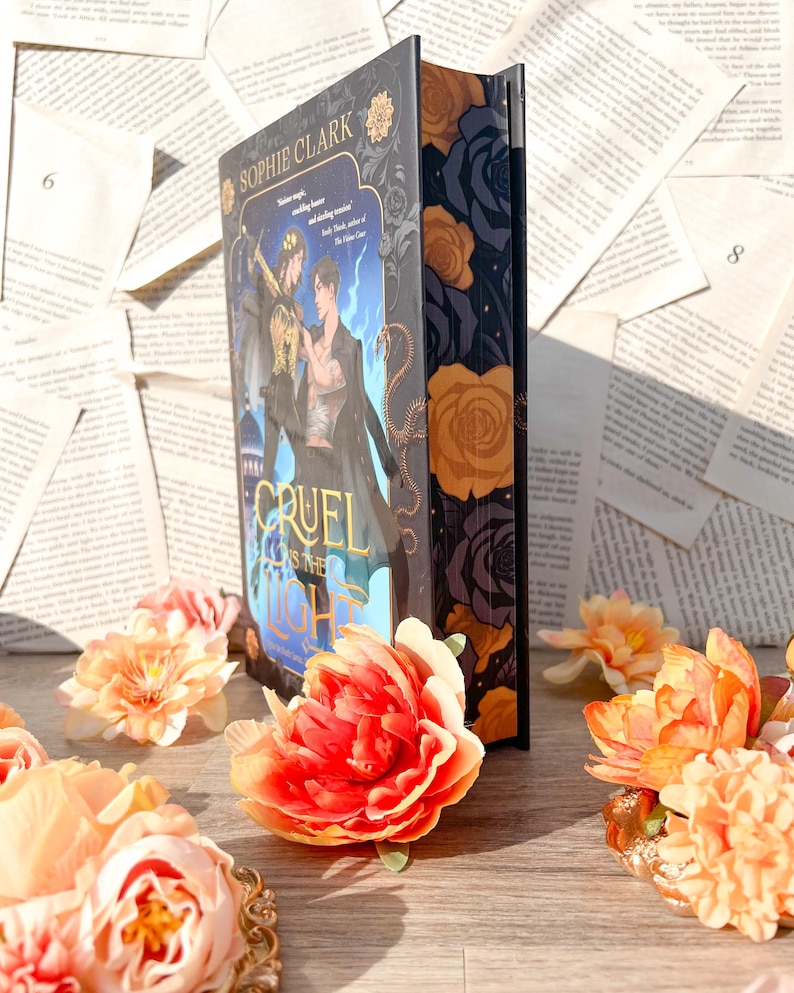May include: A hardcover book with a dark blue cover and gold lettering. The title of the book is "Cruel Prince" and the author is Sophie Clark. The cover features a dark blue background with a gold snake and a gold crown. The book is surrounded by pink and orange flowers.