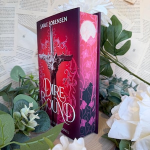 Op de afbeelding: Een hardcover boek getiteld "Dire Bound" van Sable Sorensen. De omslag is dieprood met een zilveren zwaard- en drakenontwerp. Het boek is omgeven door witte bloemen en groen, met een achtergrond van oude boekpagina's.