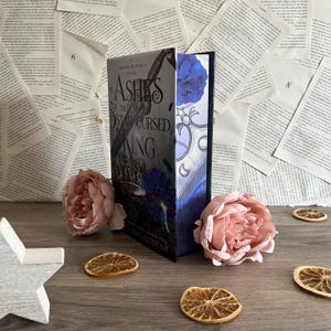 May include: A hardcover book titled "The Ashes & The Star Cursed King" with a dark cover design. The book is propped up with a floral arrangement and dried orange slices. The background features scattered book pages.