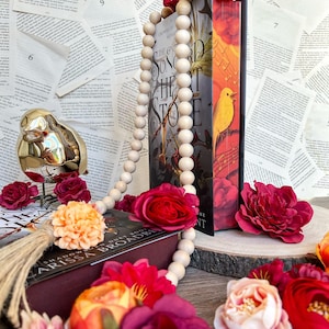 May include: A book titled "The Song of the Heart of Stone" by  A.C. Gaughen is displayed with a wooden bead garland and several red, pink, and orange silk flowers.