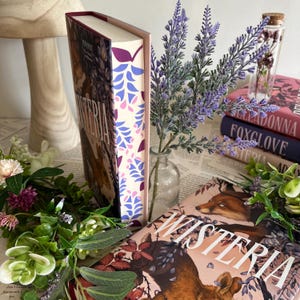 May include: A stack of books with floral and fox illustrations. The book on top is titled "Wisteria" and has a fox illustration. The spine of the book has a floral pattern in purple and blue. Lavender sprigs and other greenery surround the books.