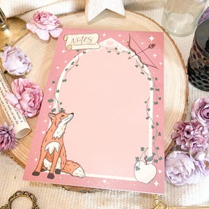 May include: A pink notepad featuring a fox illustration, bow and arrow, and apple. The notepad has the word "Notes" at the top. The design is framed with a green vine and white stars. Decorative flowers and a wooden star surround the notepad.