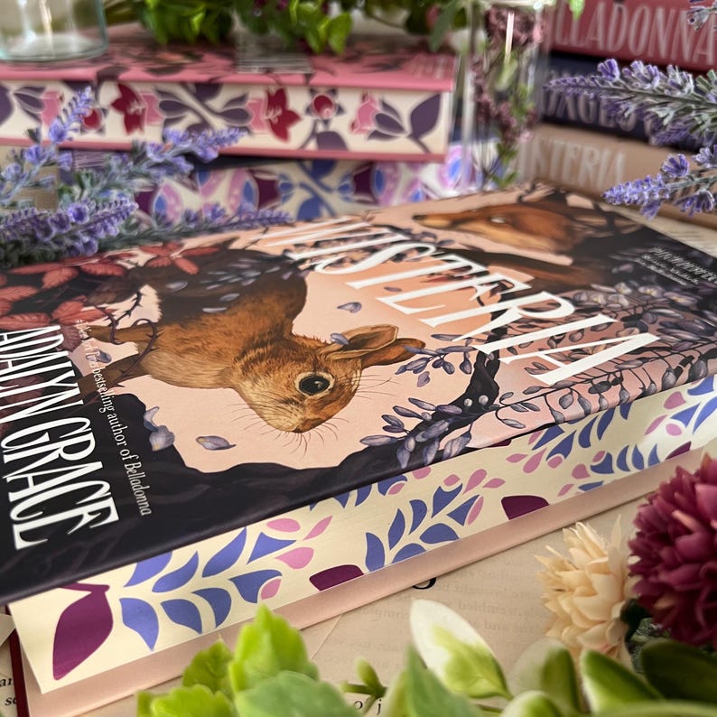 May include: A stack of books with floral and botanical designs. The top book features a brown rabbit illustration and the title "Mysterium" by Adalyn Grace. Other books have pink and purple floral patterns.