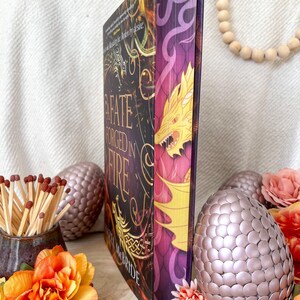 Könnte beinhalten: Ein Hardcover-Buch mit dem Titel "A Fate Forged in Fire" mit einer Drachenillustration. Das Buch hat einen dunklen Einband mit goldenen und lila Akzenten. Dekorative eiförmige Objekte und Blumen umgeben das Buch.