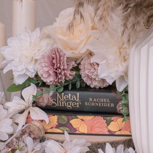 May include: Two stacked books with floral arrangements. The top book is black with the text Metal Slinger by Rachel Schneider. The bottom book features a yellow and orange butterfly and floral pattern. White and dusty pink artificial flowers surround.