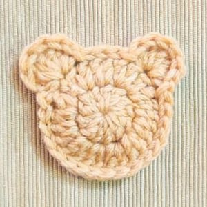 Può includere: Un viso di orso all'uncinetto di colore beige chiaro. Il viso dell'orso è rotondo con due piccole orecchie sulla parte superiore. Le cuciture all'uncinetto sono visibili, creando un aspetto strutturato. L'orso è appoggiato su un tessuto beige chiaro a righe verticali.