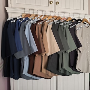 May include: A row of 14 shirts hanging on hangers, showcasing a variety of colours, including navy, dark blue, light blue, grey, dark brown, dark tan, mid tan, light tan, dark green, mid green, light green, dark grey, and light grey.