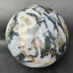 May include: A polished agate sphere with a swirling pattern of white, gray, black, and green. The sphere is smooth and has a natural, organic look.