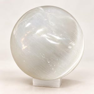 May include: A polished, spherical selenite crystal displaying a translucent, milky white appearance with fibrous, chatoyant internal textures. The sphere rests securely upon a small, rectangular, white selenite base.