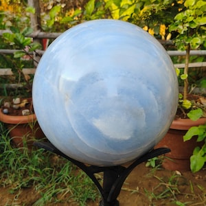 May include: A large, light blue sphere with white marbling, resting on a black metal stand. The sphere has a smooth, polished surface and is displayed outdoors with greenery in the background. The sphere is a decorative garden accent.