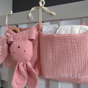 May include: A pink fabric crib organizer with three pockets. The organizer has a stuffed bunny toy with the name "Olivia" embroidered on it. The pocket on the right has the name "Ophelia" embroidered on it.