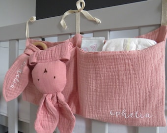 May include: A dusty pink muslin hanging organizer with two pockets, featuring the name Ophelia embroidered in white script. The left pocket holds a plush rabbit toy with long ears and a wooden ring, while the right pocket contains a diaper and wipes.