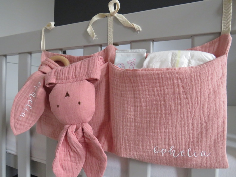 May include: A pink fabric crib organizer with three pockets. The organizer has a stuffed bunny toy with the name "Olivia" embroidered on it. The pocket on the right has the name "Ophelia" embroidered on it.