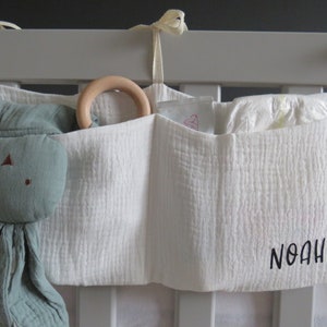 May include: White fabric crib organizer with three pockets. The organizer has a wooden ring toy in the top pocket, a green stuffed animal in the left pocket, and a diaper in the right pocket. The organizer is personalized with the name "Noah" in black embroidery.