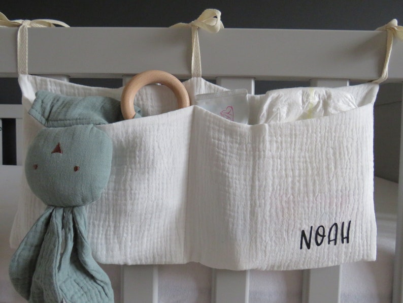 May include: White fabric crib organizer with three pockets. The organizer has a wooden ring toy in the top pocket, a green stuffed animal in the left pocket, and a diaper in the right pocket. The organizer is personalized with the name "Noah" in black embroidery.