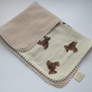 Puede incluir: Una manta de bebé plegada, en tonos beige y crema. Presenta un patrón de ositos de peluche marrones y está adornada con pompones blancos. La manta está hecha de una tela suave y texturizada. Ideal para un bebé.