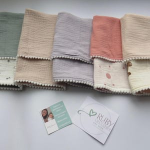 May include: A collection of folded muslin baby blankets in various colors, including green, beige, gray, coral, and cream. Each blanket features a white pom-pom trim and a patterned bottom with stars, dots, or a teddy bear. Two cards are also visible.