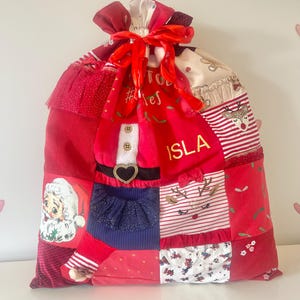 May include: A large, red Christmas gift bag with a patchwork design. The bag features a Santa Claus graphic, reindeer, and the name "ISLA". It is tied with a red ribbon.