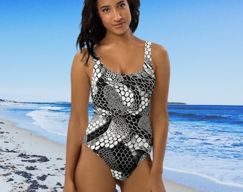 May include: A black and white swimsuit with a geometric pattern. The swimsuit has a scoop neck and a low back. The model is wearing the swimsuit on a beach.