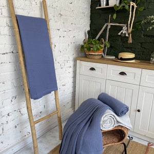 May include: Two blue waffle weave blankets, one folded and placed on a wooden ladder, the other folded and placed on a wicker stool with a white blanket underneath. The stool is on a woven rug with black and white stripes.