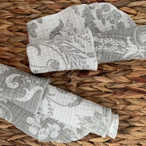 May include: Two gray and white patterned fabric squares folded and stacked on a woven brown surface.