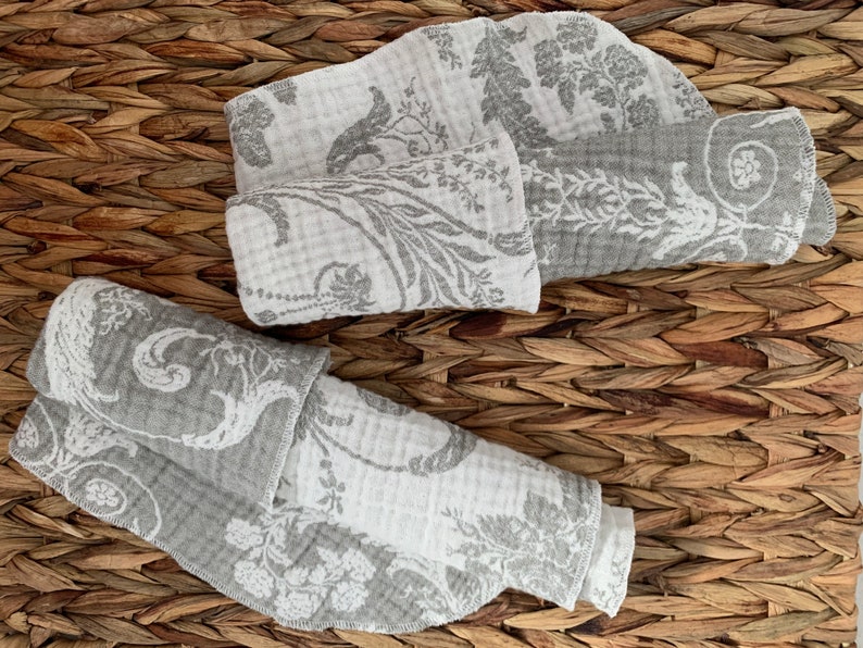 May include: Two gray and white patterned fabric squares folded and stacked on a woven brown surface.