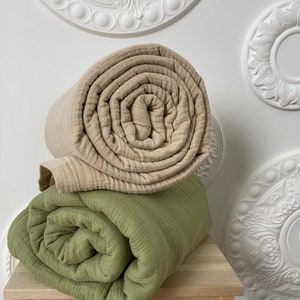May include: Two rolled up blankets, one beige and one green, are stacked on a wooden stool. The stool is in front of a white wall with decorative white ceiling medallions.