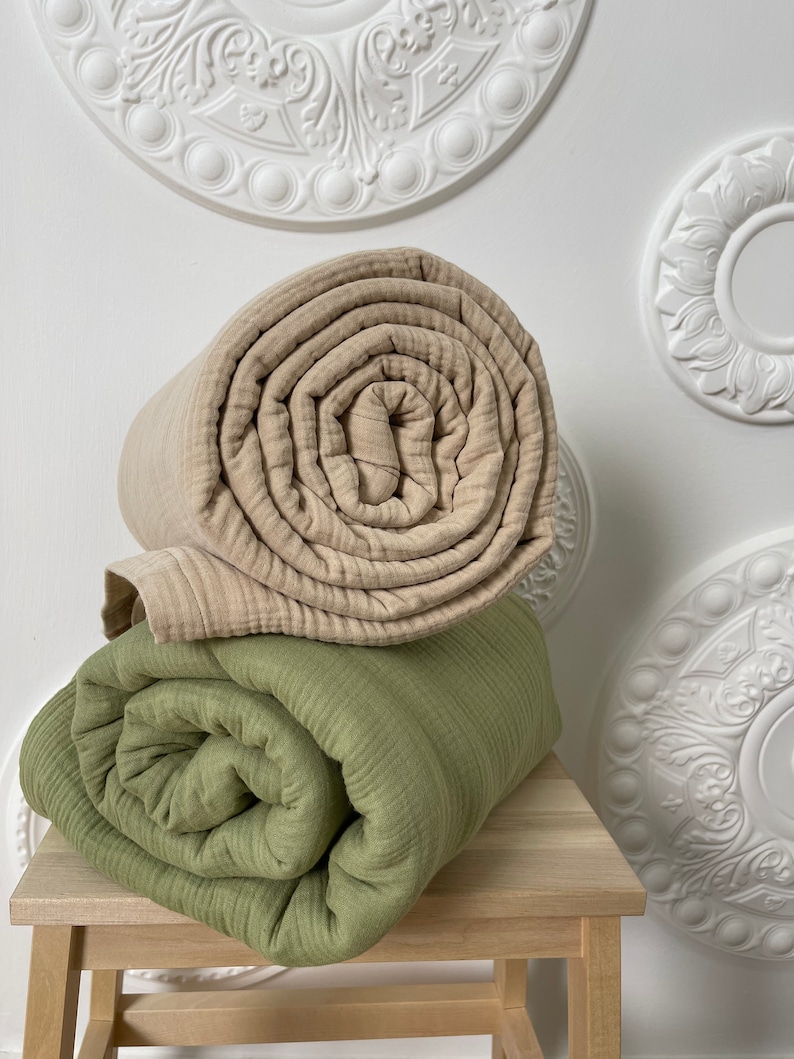 May include: Two rolled up blankets, one beige and one green, are stacked on a wooden stool. The stool is in front of a white wall with decorative white ceiling medallions.