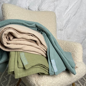 May include: A stack of three muslin baby blankets in light blue, beige, and green. The blankets are folded and arranged on a white armchair. Each blanket has a small label with the word "Little" printed on it.