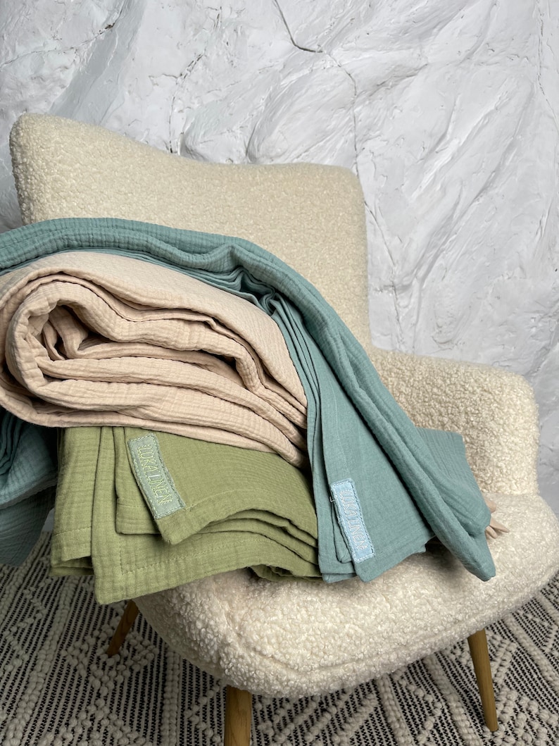 May include: A stack of three muslin baby blankets in light blue, beige, and green. The blankets are folded and arranged on a white armchair. Each blanket has a small label with the word "Little" printed on it.