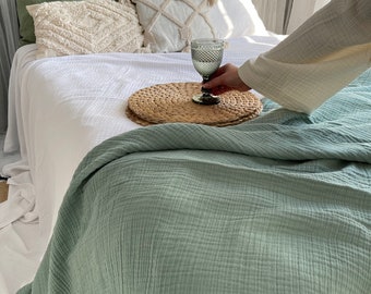 May include: A sage green waffle weave throw blanket is draped over a white bed with a woven wicker tray and a clear glass with a stem.