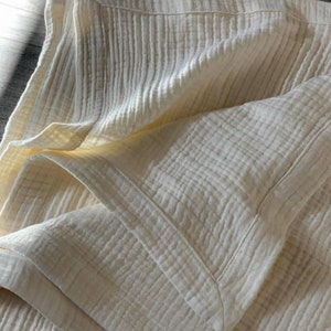 May include: A white cotton muslin blanket with a textured waffle weave. The blanket is folded in half and the edges are sewn with a white thread.