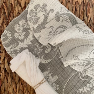 May include: A set of two kitchen towels, one white and one gray with a floral pattern. The gray towel is folded and the white towel is rolled up and tied with twine.