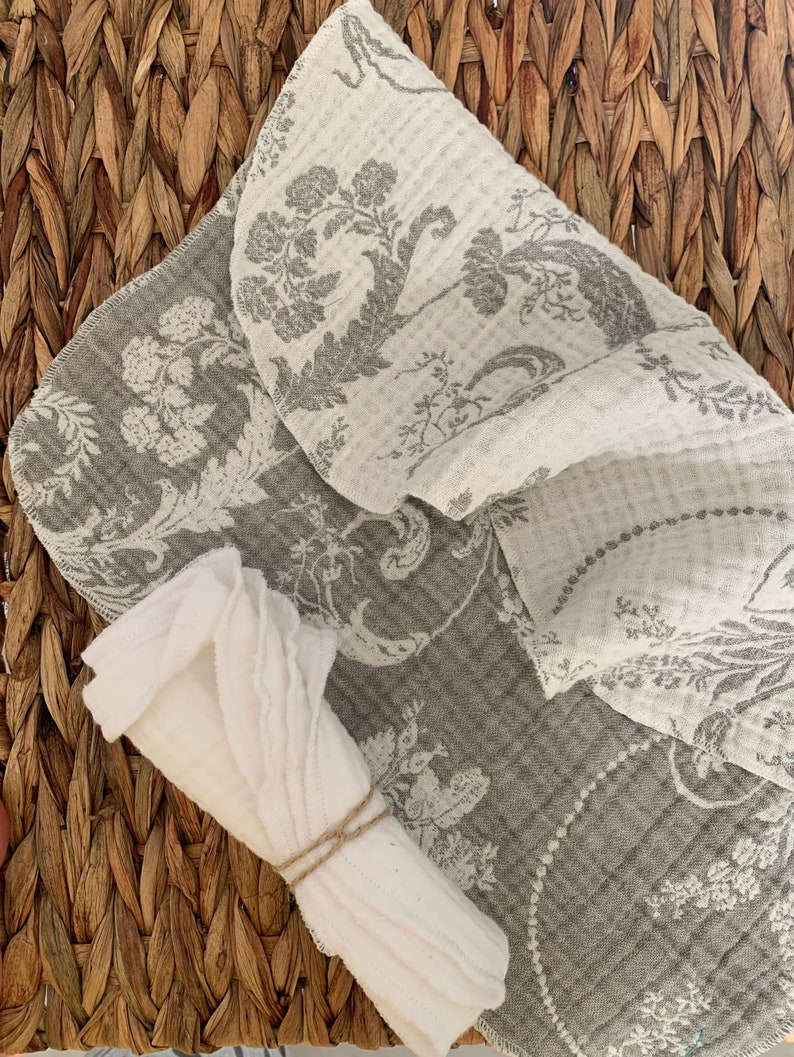 May include: A set of two kitchen towels, one white and one gray with a floral pattern. The gray towel is folded and the white towel is rolled up and tied with twine.