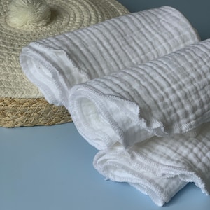 May include: A stack of white cotton muslin cloths with a decorative stitched edge. The cloths are folded and rolled up.