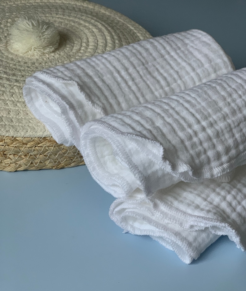 May include: A stack of white cotton muslin cloths with a decorative stitched edge. The cloths are folded and rolled up.