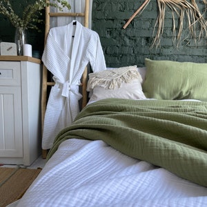 May include: A white waffle weave robe hanging on a bamboo ladder, with a green waffle weave throw blanket and white waffle weave bedding on a bed with a green wall and a large dried palm leaf hanging above the bed.