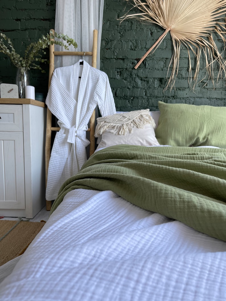 May include: A white waffle weave robe hanging on a bamboo ladder, with a green waffle weave throw blanket and white waffle weave bedding on a bed with a green wall and a large dried palm leaf hanging above the bed.