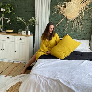 Pu&ograve; includere: Una donna in una veste gialla &egrave; seduta su un letto bianco con un cuscino giallo. Il letto ha una coperta bianca e un copriletto nero. La stanza ha un muro di mattoni verdi, una tenda bianca e una grande foglia di palma secca appesa al muro.