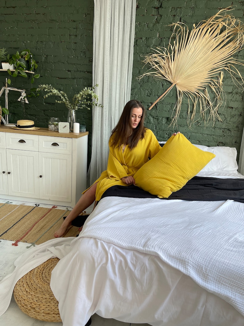 Pu&ograve; includere: Una donna in una veste gialla &egrave; seduta su un letto bianco con un cuscino giallo. Il letto ha una coperta bianca e un copriletto nero. La stanza ha un muro di mattoni verdi, una tenda bianca e una grande foglia di palma secca appesa al muro.