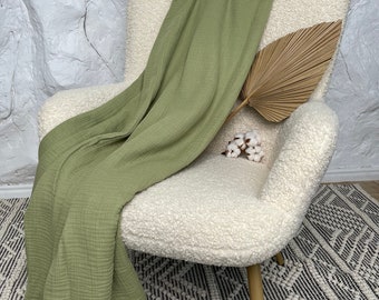 May include: A sage green waffle weave throw blanket draped over a white faux fur armchair with a dried palm leaf and cotton balls on the seat.