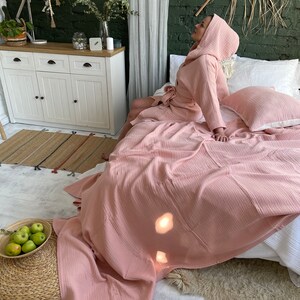 Pu&ograve; includere: Una vestaglia rosa in tessuto a nido d'ape con cappuccio, indossata da una persona seduta su un letto con biancheria da letto bianca e una coperta in tessuto a nido d'ape rosa. Il letto si trova in una stanza con una cassettiera bianca e un muro di mattoni verdi.