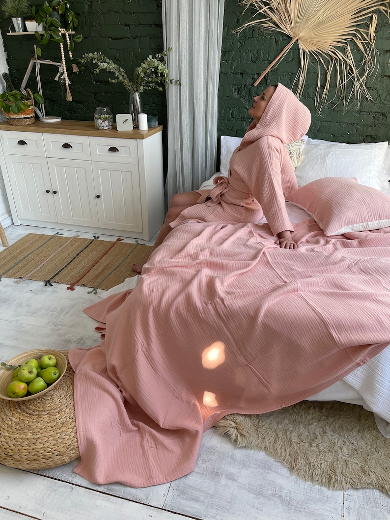 Pu&ograve; includere: Una vestaglia rosa in tessuto a nido d'ape con cappuccio, indossata da una persona seduta su un letto con biancheria da letto bianca e una coperta in tessuto a nido d'ape rosa. Il letto si trova in una stanza con una cassettiera bianca e un muro di mattoni verdi.