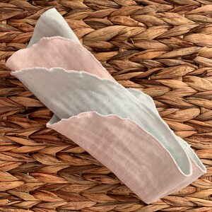 May include: Two folded cotton cloths, one light pink and one light blue, with scalloped edges. The cloths are folded and placed on a woven brown surface.